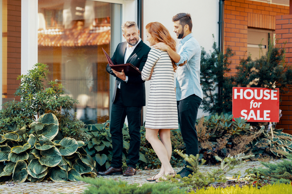 man in suit talks to couple in front of building with "house for sale" sign